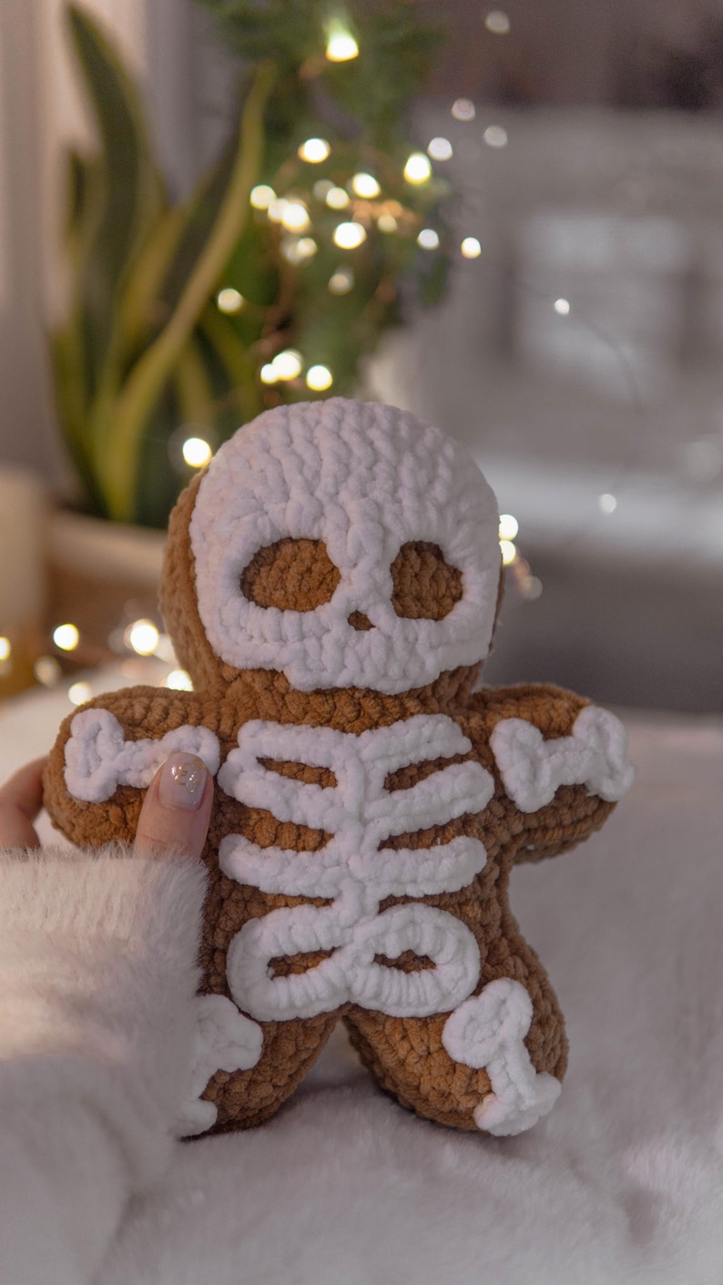 May include: A brown crocheted gingerbread man with a white skull face and skeleton design. The plush toy is handmade and has a soft, textured appearance. The background is blurred, with hints of greenery and fairy lights.