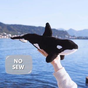 Puede incluir: Un peluche de orca tejido a ganchillo en blanco y negro con aleta dorsal, aletas laterales y aleta caudal. El peluche tiene una mancha blanca cerca del ojo y la parte inferior blanca. Se ve un gráfico gris con el texto NO SEW.