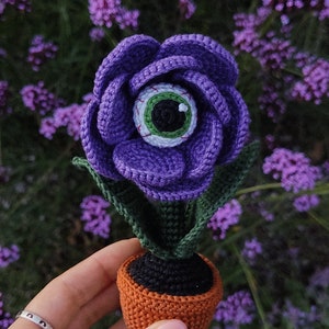 Puede incluir: Una flor de color morado tejida a crochet con un ojo verde en el centro, en maceta de color naranja. La flor est&aacute; sentada en una mano sobre un fondo de flores moradas.