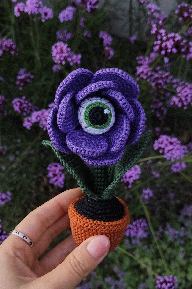 Puede incluir: Una flor de color morado tejida a crochet con un ojo verde en el centro, en maceta de color naranja. La flor est&aacute; sentada en una mano sobre un fondo de flores moradas.