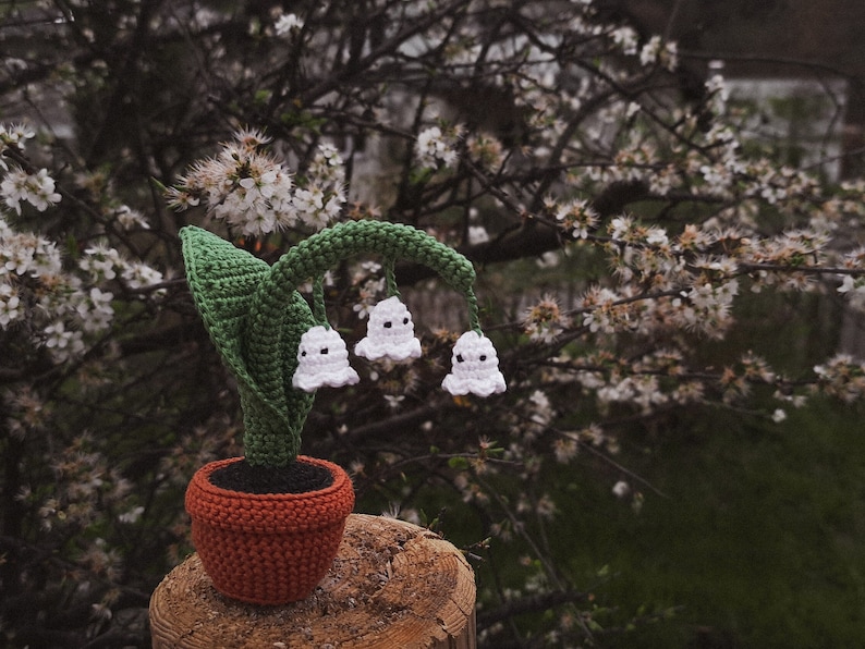 May include: A crocheted lily of the valley plant with white flowers in a brown pot. The plant is sitting on a wooden surface.