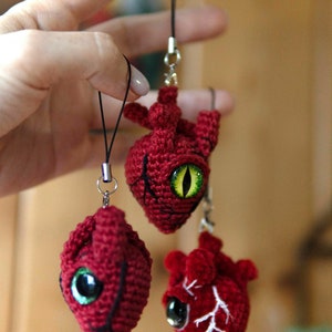 Könnte beinhalten: Drei gehäkelte rote herzförmige Schlüsselanhänger mit grünen Augen. Jeder Schlüsselanhänger hat ein anderes Design, einer mit einem aufgenähten Auge, einer mit einem bemalten Auge und einer mit einem detaillierten anatomischen Design.