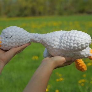 May include: A handmade white crochet goose plush toy with orange accents on the beak and feet. The soft toy is held up against a blurred green background, suggesting an outdoor setting. The goose has closed eyes, giving it a sleepy appearance.