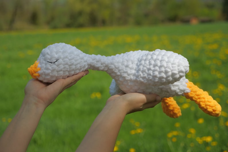 May include: A handmade white crochet goose plush toy with orange accents on the beak and feet. The soft toy is held up against a blurred green background, suggesting an outdoor setting. The goose has closed eyes, giving it a sleepy appearance.