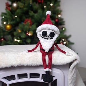 May include: A crocheted skeleton Santa Claus doll wearing a red hat and suit with a white beard. The doll is sitting on a white fluffy surface.