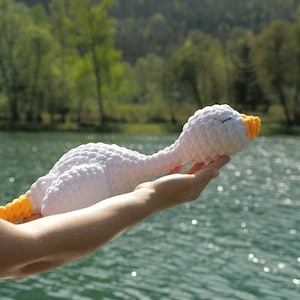 May include: A handmade white crochet goose with a yellow beak and neck. The plush toy is held up against a backdrop of a lake and trees. The goose has closed eyes, suggesting it is sleeping.