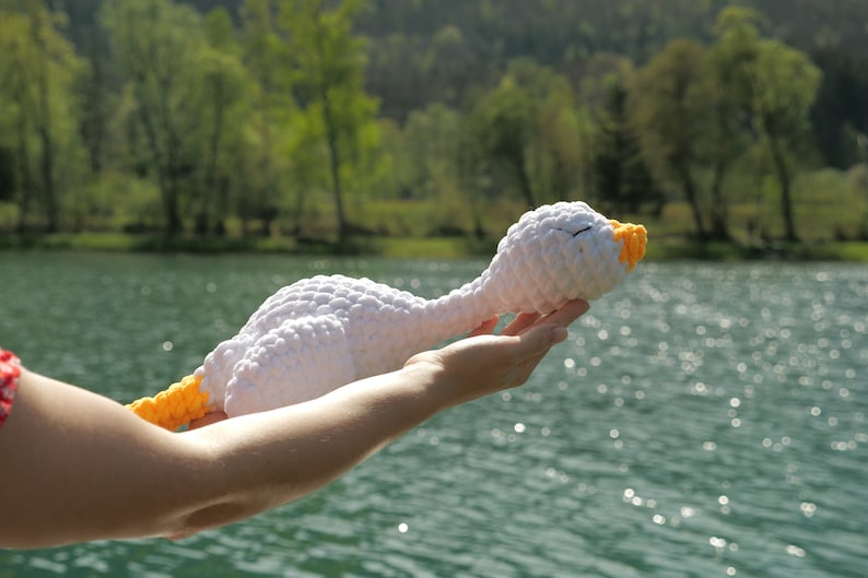 May include: A handmade white crochet goose with a yellow beak and neck. The plush toy is held up against a backdrop of a lake and trees. The goose has closed eyes, suggesting it is sleeping.