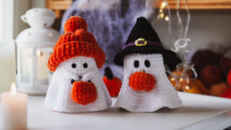K&ouml;nnte beinhalten: Zwei geh&auml;kelte Geister-Ornamente, eines mit einem schwarzen Hexenhut mit einem violetten Band und das andere mit einer orangefarbenen Strickm&uuml;tze. Beide Geister haben orangefarbene geh&auml;kelte Nasen und schwarze Augen.