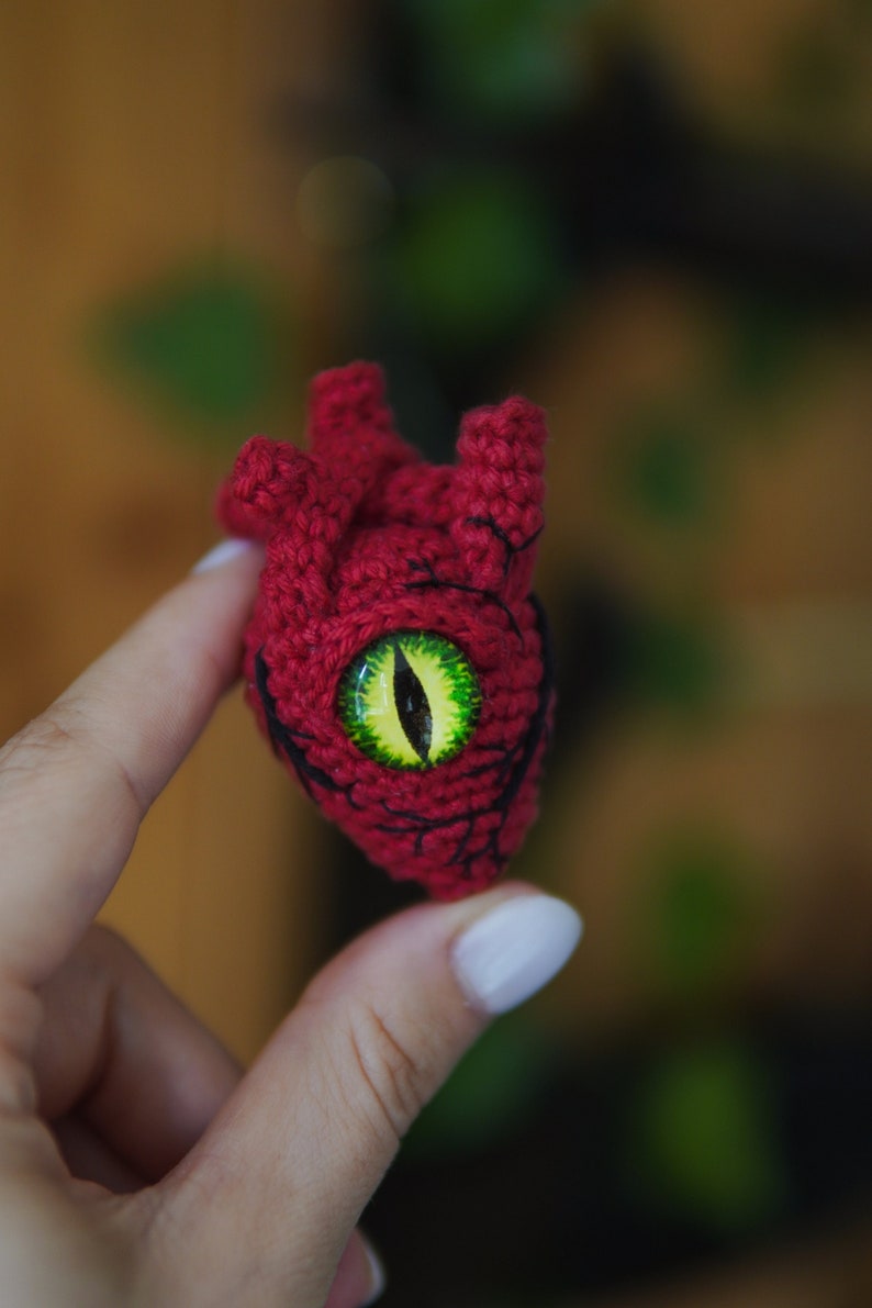 May include: A crocheted heart with a green eye, the heart is red and has a textured surface.