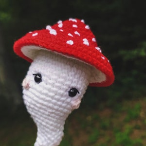 May include: A crocheted mushroom toy with a red and white cap and a white body. The mushroom has black eyes and a pink nose.