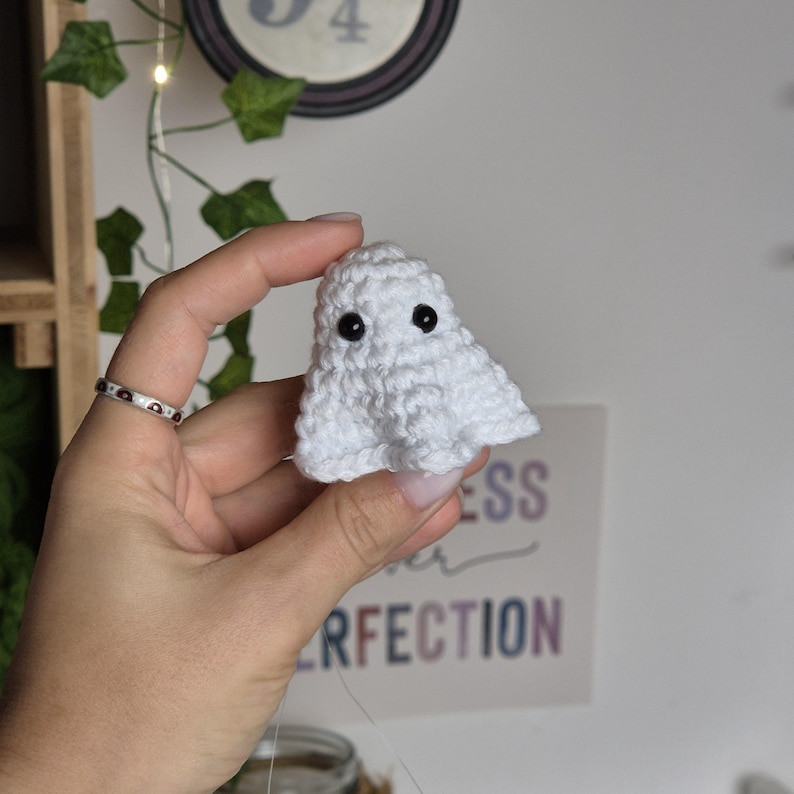 Puede incluir: Un peque&ntilde;o fantasma blanco de ganchillo hecho a mano con ojos de bot&oacute;n negros. El fantasma se sostiene en una mano, mostrando su apariencia texturizada y esponjosa. El fondo incluye un reloj decorativo y un letrero con la palabra "PERFECTION".