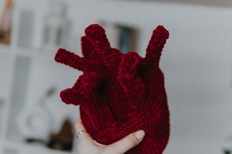 May include: A crocheted plush heart shaped toy in a deep red color. The heart has a textured surface and is made with a soft, fuzzy yarn.