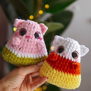 Op de afbeelding: Twee gehaakte pluche speelgoedjes in de vorm van candy corn en watermeloen. Het candy corn speelgoed is wit, oranje en geel. Het watermeloen speelgoed is roze, groen en wit.