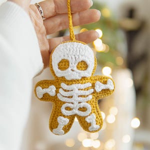 May include: A crocheted gingerbread man ornament in a skeleton design. The ornament is yellow with white crochet details for the skull and skeleton. It has a yellow loop for hanging.