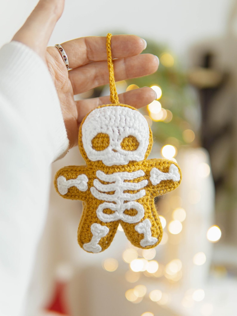 May include: A crocheted gingerbread man ornament in a skeleton design. The ornament is yellow with white crochet details for the skull and skeleton. It has a yellow loop for hanging.