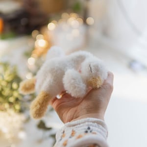 May include: A small, handmade white plush bunny with tan accents on its ears and feet. The bunny is lying on its side, held in a person's hand. The bunny's fur appears soft and fluffy. The background is blurred with soft lighting.