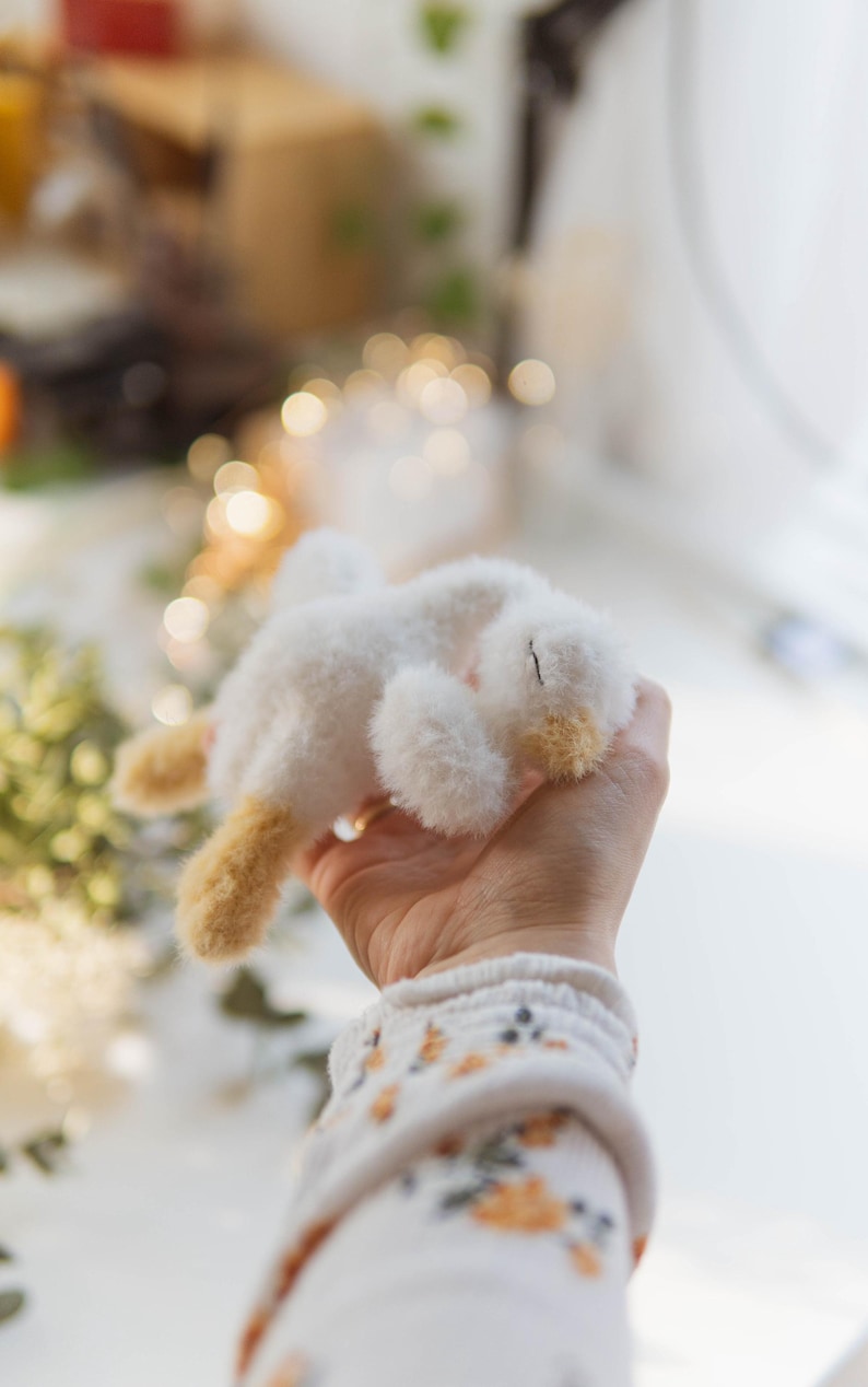 May include: A small, handmade white plush bunny with tan accents on its ears and feet. The bunny is lying on its side, held in a person's hand. The bunny's fur appears soft and fluffy. The background is blurred with soft lighting.