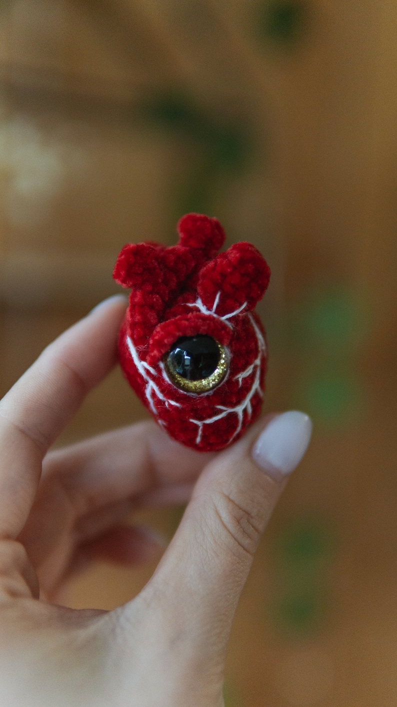 May include: A crocheted red heart with white stitching and a single black eye with gold glitter.