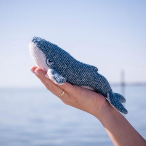 May include: A crocheted whale toy in shades of blue and white. The toy is held in a hand, highlighting its detailed crochet work. The whale's design is visible against a backdrop of the sea and sky, with a textured finish.