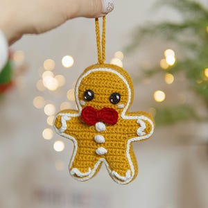 May include: A crocheted gingerbread man Christmas ornament. The ornament is yellow with white trim, black eyes, and a red bow tie. It is suspended by a matching yellow string. The background is blurred with bokeh lights.