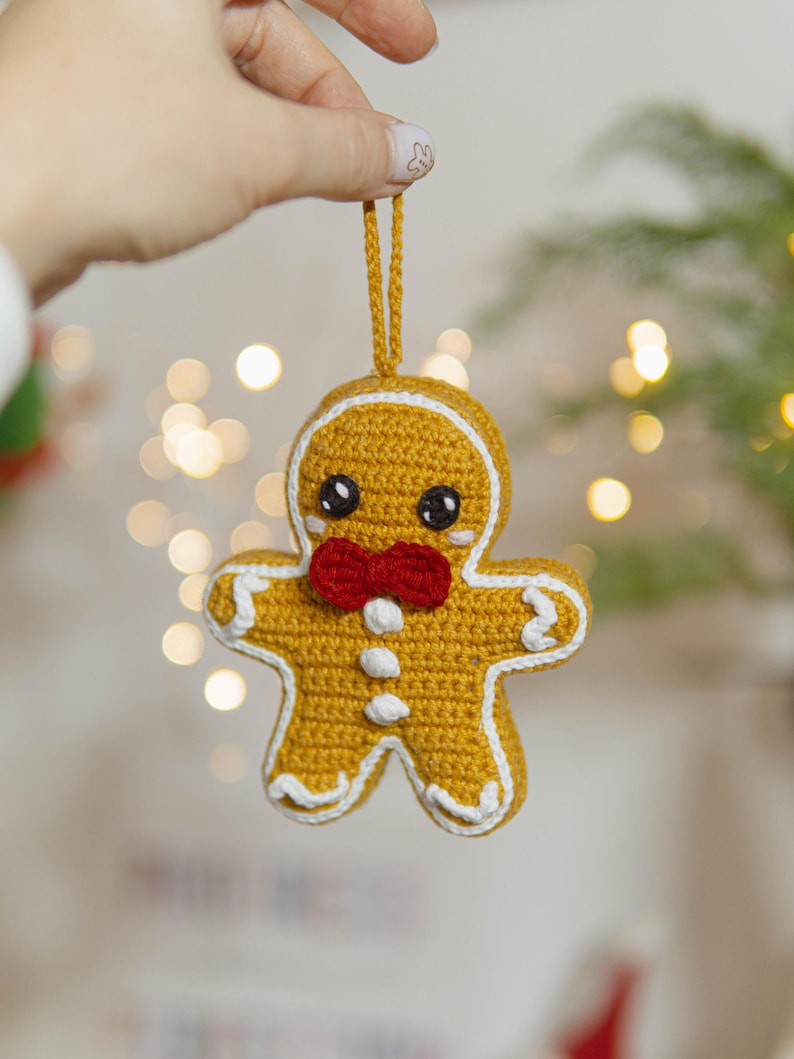May include: A crocheted gingerbread man Christmas ornament. The ornament is yellow with white trim, black eyes, and a red bow tie. It is suspended by a matching yellow string. The background is blurred with bokeh lights.