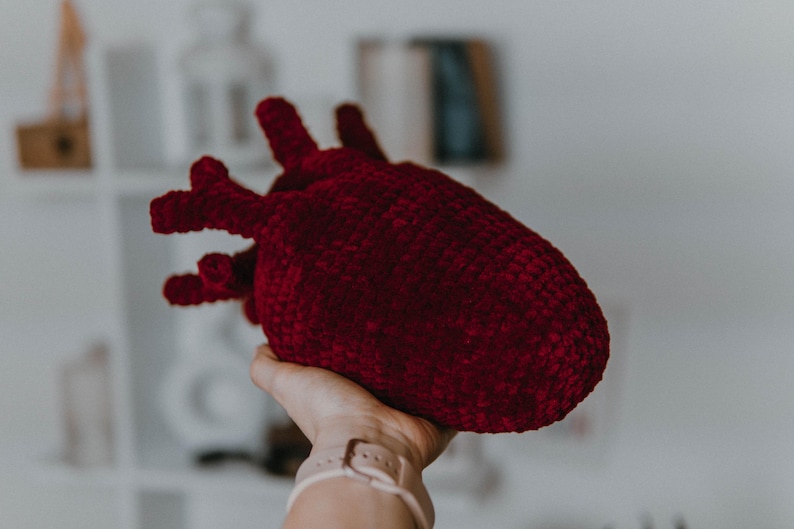 May include: A crocheted plush heart shaped toy in a deep red color. The heart is being held in a person's hand.
