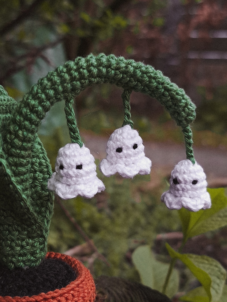 May include: A crocheted lily of the valley plant with white flowers and green leaves in a brown pot.