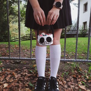 K&ouml;nnte beinhalten: Eine Person tr&auml;gt einen schwarzen Rock, wei&szlig;e Socken und schwarze Sneaker und h&auml;lt einen wei&szlig;en geh&auml;kelten Korb mit einem Jack Skellington Gesicht. Der Korb ist mit bunten S&uuml;&szlig;igkeiten gef&uuml;llt.