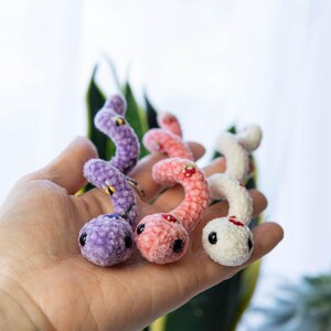K&ouml;nnte beinhalten: Drei geh&auml;kelte Wurm-Pl&uuml;schtiere in verschiedenen Farben: Lila, Rosa und Wei&szlig;. Jeder Wurm hat gestickte Details, darunter Augen, einen Mund und ein kleines Design auf dem R&uuml;cken.