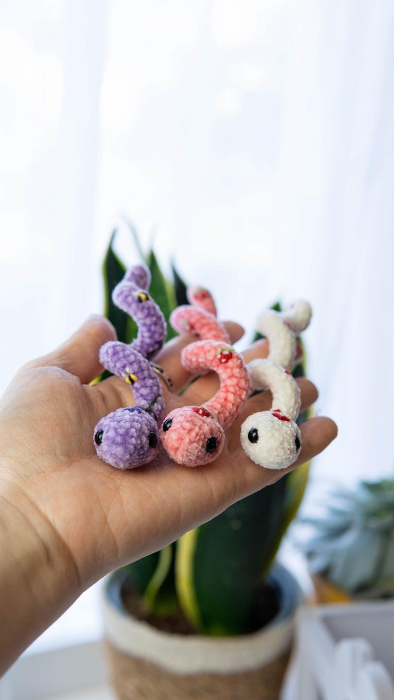 K&ouml;nnte beinhalten: Drei geh&auml;kelte Wurm-Pl&uuml;schtiere in verschiedenen Farben: Lila, Rosa und Wei&szlig;. Jeder Wurm hat gestickte Details, darunter Augen, einen Mund und ein kleines Design auf dem R&uuml;cken.