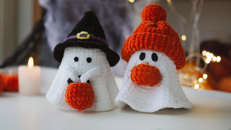K&ouml;nnte beinhalten: Zwei geh&auml;kelte Geister-Ornamente, eines mit einem schwarzen Hexenhut und einem orangefarbenen K&uuml;rbis, das andere mit einer orangefarbenen Strickm&uuml;tze mit Bommel.