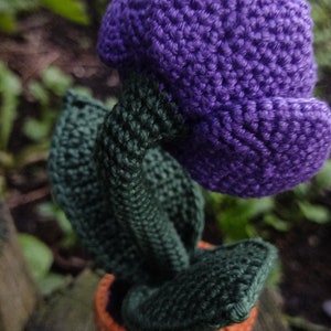Puede incluir: Una flor de color morado tejida a crochet con hojas verdes y una maceta naranja. La flor est&aacute; hecha de hilo y es una artesan&iacute;a hecha a mano.