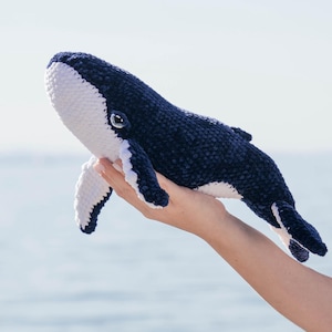 K&ouml;nnte beinhalten: Ein Kuscheltier in Form eines Wals, in Marineblau und Wei&szlig;. Der Wal wird vor dem Meer gehalten. Das Spielzeug hat eine abgerundete Form mit wei&szlig;en Details an Bauch und Flossen. Das Auge des Wals ist sichtbar.