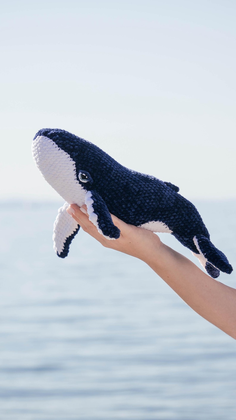 K&ouml;nnte beinhalten: Ein Kuscheltier in Form eines Wals, in Marineblau und Wei&szlig;. Der Wal wird vor dem Meer gehalten. Das Spielzeug hat eine abgerundete Form mit wei&szlig;en Details an Bauch und Flossen. Das Auge des Wals ist sichtbar.