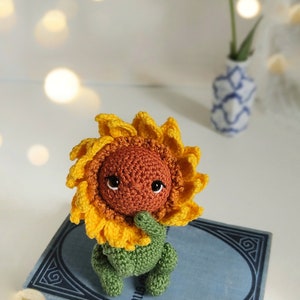 K&ouml;nnte beinhalten: Ein geh&auml;keltes Sonnenblumen-Amigurumi-Spielzeug mit braunem Gesicht und gr&uuml;nem Stiel, das auf einem blauen Buch mit goldenen Buchstaben sitzt.