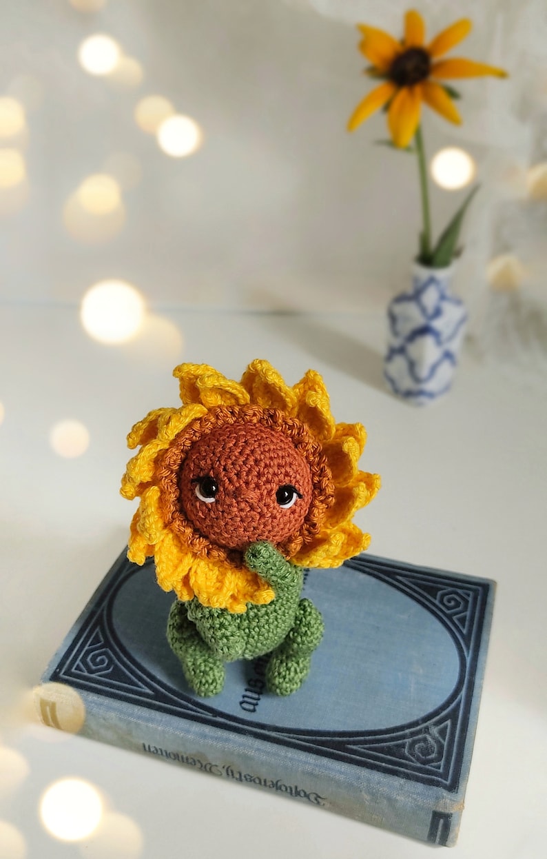 K&ouml;nnte beinhalten: Ein geh&auml;keltes Sonnenblumen-Amigurumi-Spielzeug mit braunem Gesicht und gr&uuml;nem Stiel, das auf einem blauen Buch mit goldenen Buchstaben sitzt.