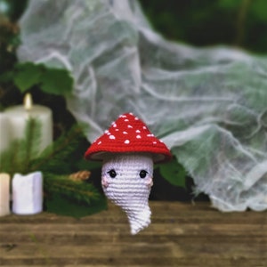 May include: A white crocheted ghost wearing a red and white crocheted mushroom hat. The ghost is sitting on a wooden surface with a white cloth in the background.