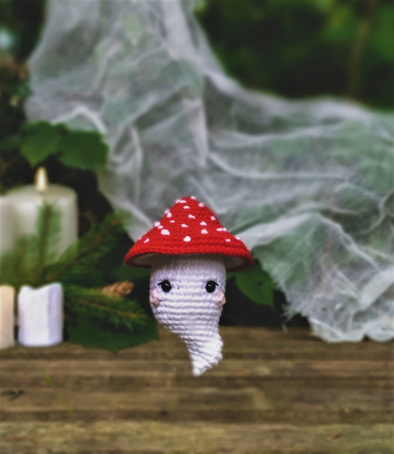 May include: A white crocheted ghost wearing a red and white crocheted mushroom hat. The ghost is sitting on a wooden surface with a white cloth in the background.