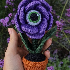 Puede incluir: Una flor de color morado tejida a crochet con un ojo verde en el centro, en maceta de color naranja. La flor se sostiene en una mano sobre un fondo de flores moradas.