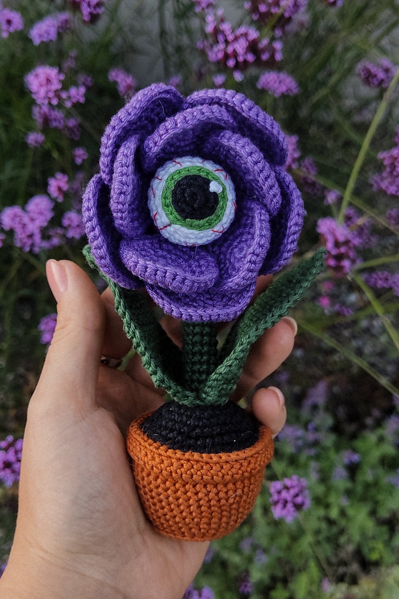 Puede incluir: Una flor de color morado tejida a crochet con un ojo verde en el centro, en maceta de color naranja. La flor se sostiene en una mano sobre un fondo de flores moradas.
