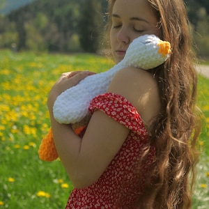 May include: A white crochet goose with orange feet and beak is held by a person wearing a red floral dress. The goose is soft and plush, with closed eyes. The background is a field of yellow flowers and green grass.