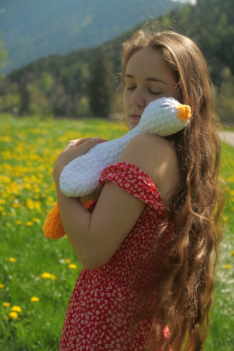 May include: A white crochet goose with orange feet and beak is held by a person wearing a red floral dress. The goose is soft and plush, with closed eyes. The background is a field of yellow flowers and green grass.