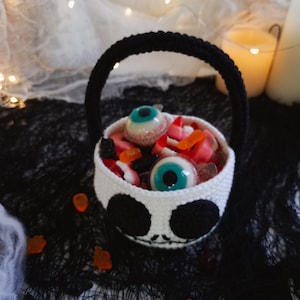 K&ouml;nnte beinhalten: Ein wei&szlig;er geh&auml;kelter Halloween-Trick-or-Treat-Korb in Form eines Sch&auml;dels mit schwarzen Augen und einem Mund. Der Korb ist gef&uuml;llt mit S&uuml;&szlig;igkeiten, darunter Gummiaugen und Gummib&auml;rchen.
