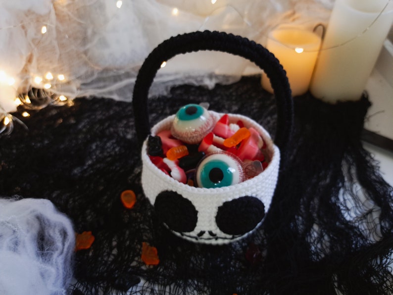 K&ouml;nnte beinhalten: Ein wei&szlig;er geh&auml;kelter Halloween-Trick-or-Treat-Korb in Form eines Sch&auml;dels mit schwarzen Augen und einem Mund. Der Korb ist gef&uuml;llt mit S&uuml;&szlig;igkeiten, darunter Gummiaugen und Gummib&auml;rchen.