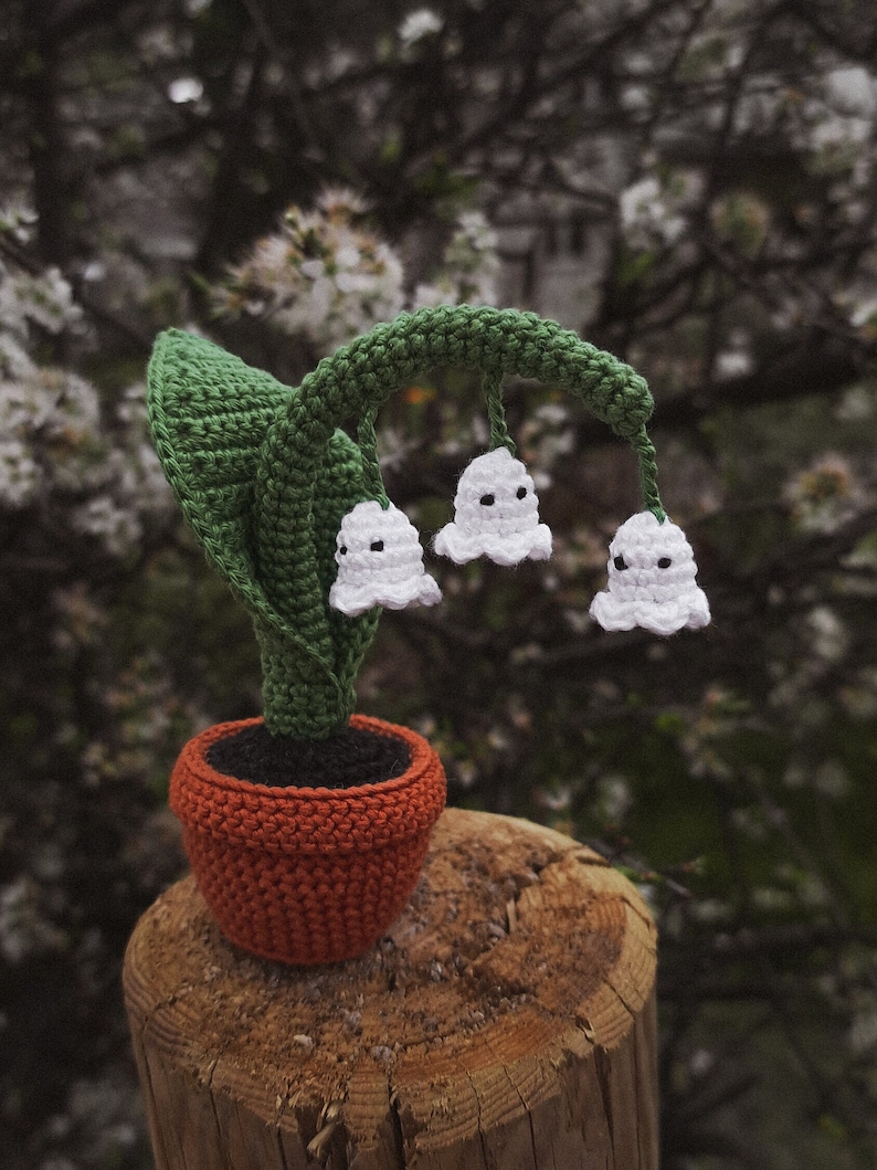 May include: A crocheted lily of the valley plant in a terracotta pot. The plant has three white flowers with a green stem and leaves.