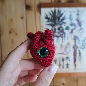 May include: A crocheted red heart with a green eye. The heart is being held in a hand.