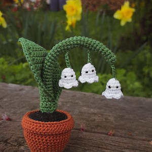 May include: A crocheted lily of the valley plant in a brown pot. The plant has white flowers with black eyes and green leaves.