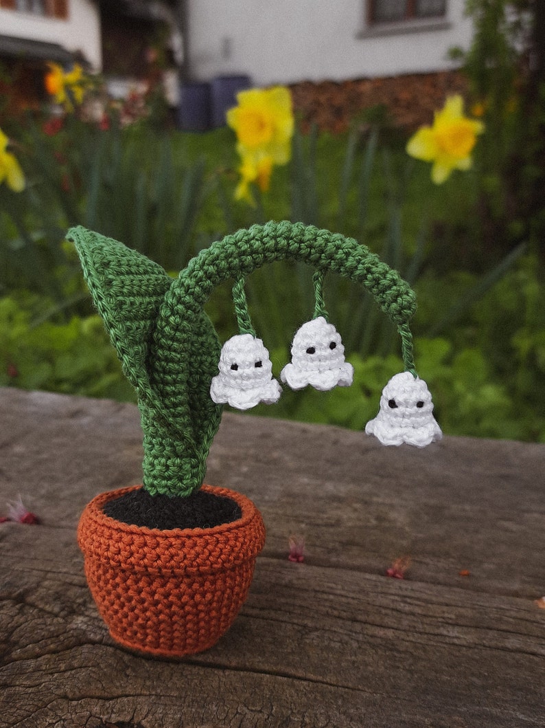May include: A crocheted lily of the valley plant in a brown pot. The plant has white flowers with black eyes and green leaves.