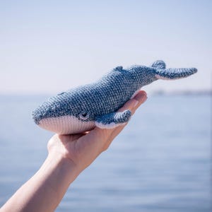 K&ouml;nnte beinhalten: Ein handgefertigtes Walspielzeug aus blauem und wei&szlig;em Garn. Der Wal wird in einer Hand gehalten, mit Meer und Himmel im Hintergrund. Der Wal hat einen hellblauen K&ouml;rper, einen wei&szlig;en Bauch und kleine Flossen.