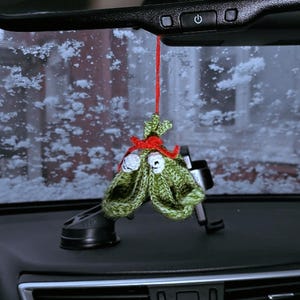 Pu&ograve; includere: Un ornamento di Natale a forma di albero di Natale in crochet verde con accenti rossi e bianchi appeso a uno specchietto retrovisore in un'auto.